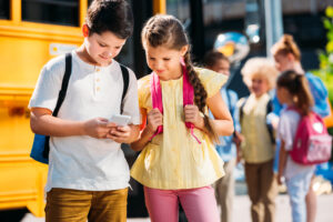 schoolgirl and schoolboy using smartphone together in front of school bus with classmates